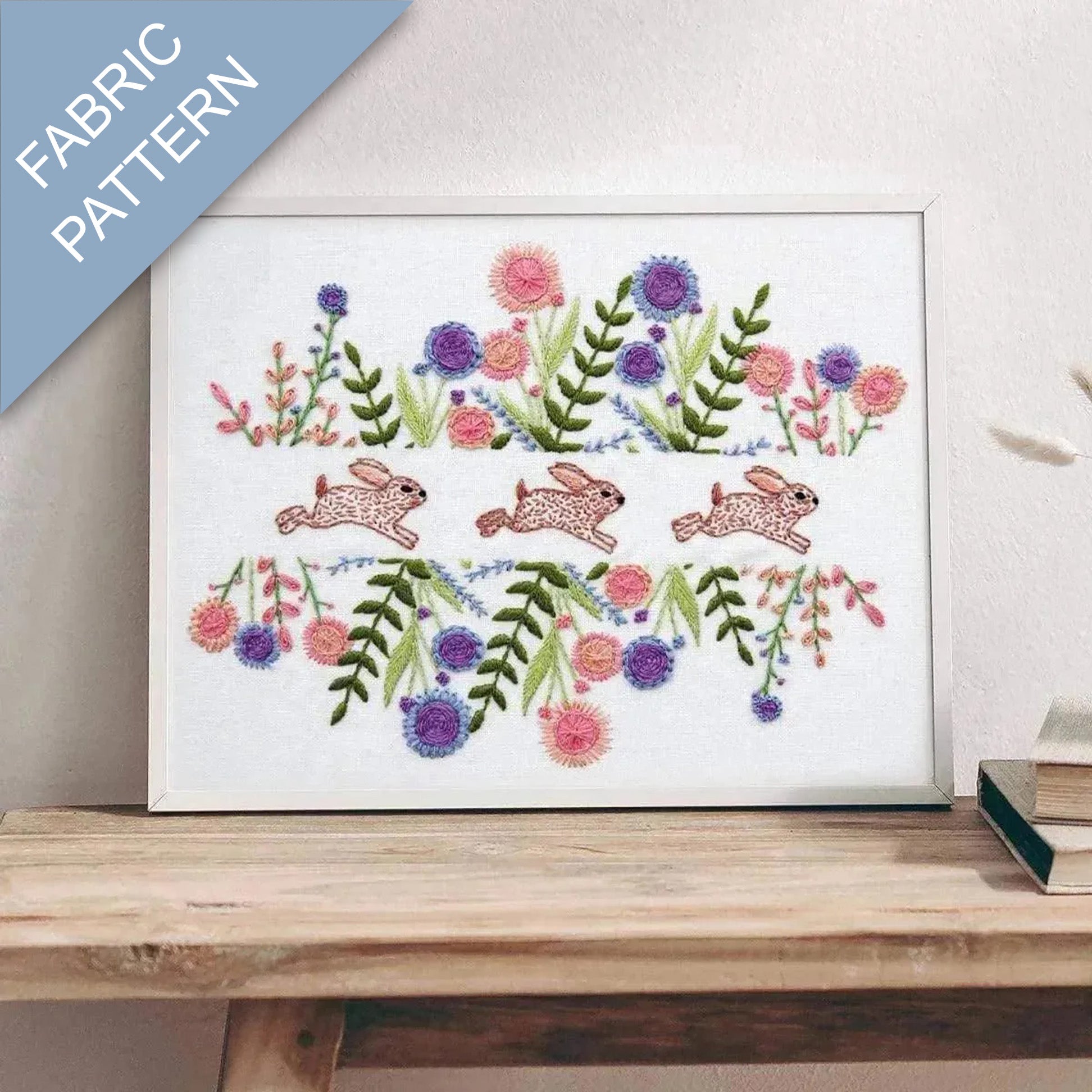 Embroidery pattern with bunnies and flowers on a white background, framed and displayed on a wooden surface.