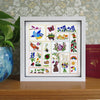 A framed square embroidery sampler featuring 15 colorful stitched animal and floral designs, displayed on a wooden shelf next to a potted plant and a red book.
