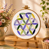 Embroidery hoop with colorful geometric pattern on a wooden stand, surrounded by flowers and stationery.