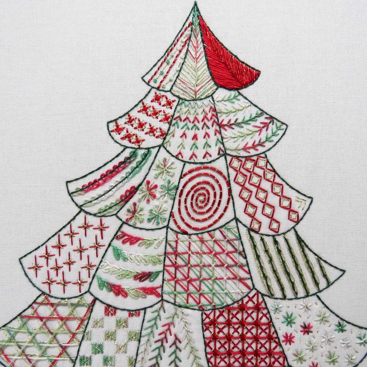 A close-up of a white fabric surface featuring a stylized Christmas tree made from stitched triangular panels in red, green, and white, with varied embroidery patterns across the tree.