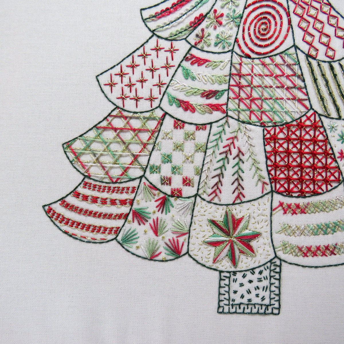 A close-up of a white fabric surface featuring a stylized Christmas tree made from stitched panels in red, green, and white with various embroidery patterns.