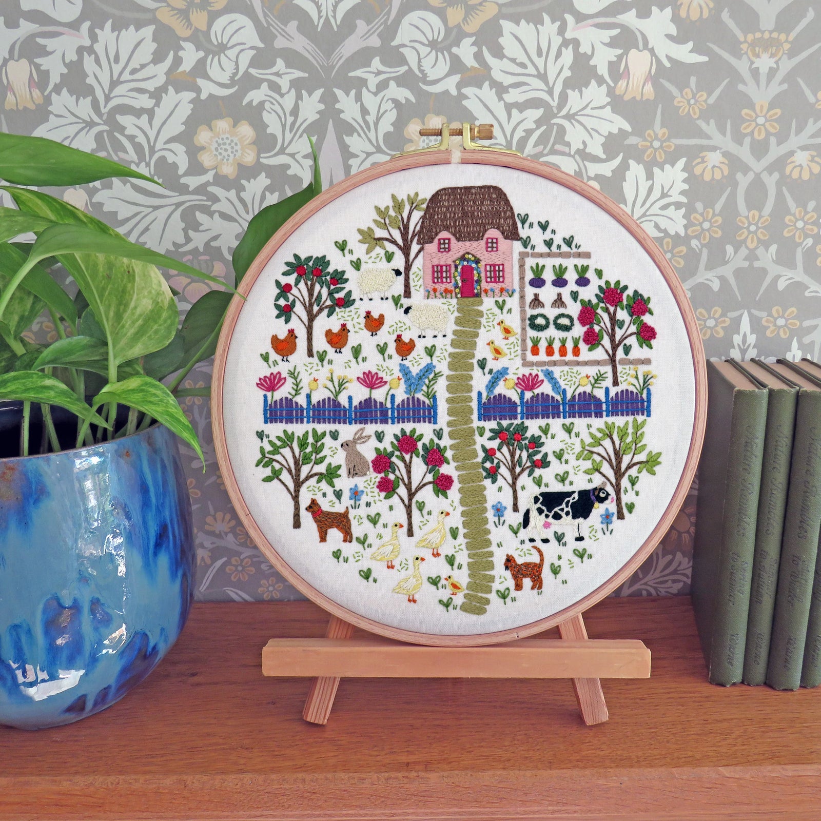 A round embroidery hoop displaying a colorful farm scene with a house, trees, chickens, cows, and a green path, mounted on a small wooden stand on a tabletop. A potted plant and books are visible in the background against patterned wallpaper.