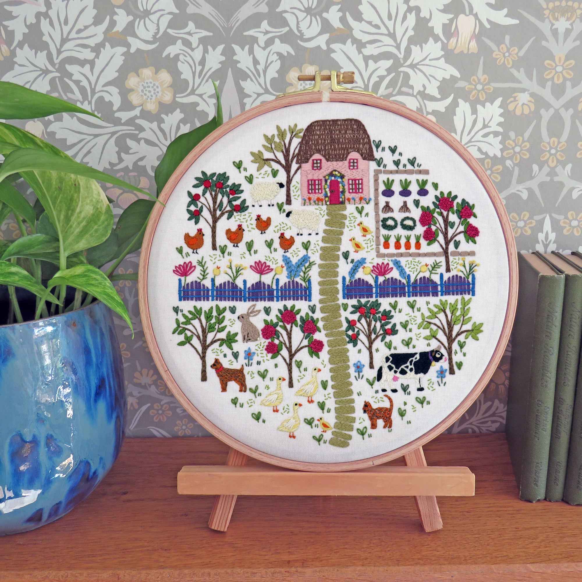 A round embroidery hoop displaying a colorful farm scene with a house, trees, chickens, cows, and a green path, mounted on a small wooden stand on a tabletop. A potted plant and books are visible in the background against patterned wallpaper.