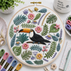 Embroidery hoop with a toucan design surrounded by flowers and leaves on a table with embroidery supplies.