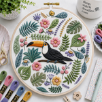 Embroidery hoop with a toucan design surrounded by flowers and leaves on a table with embroidery supplies.