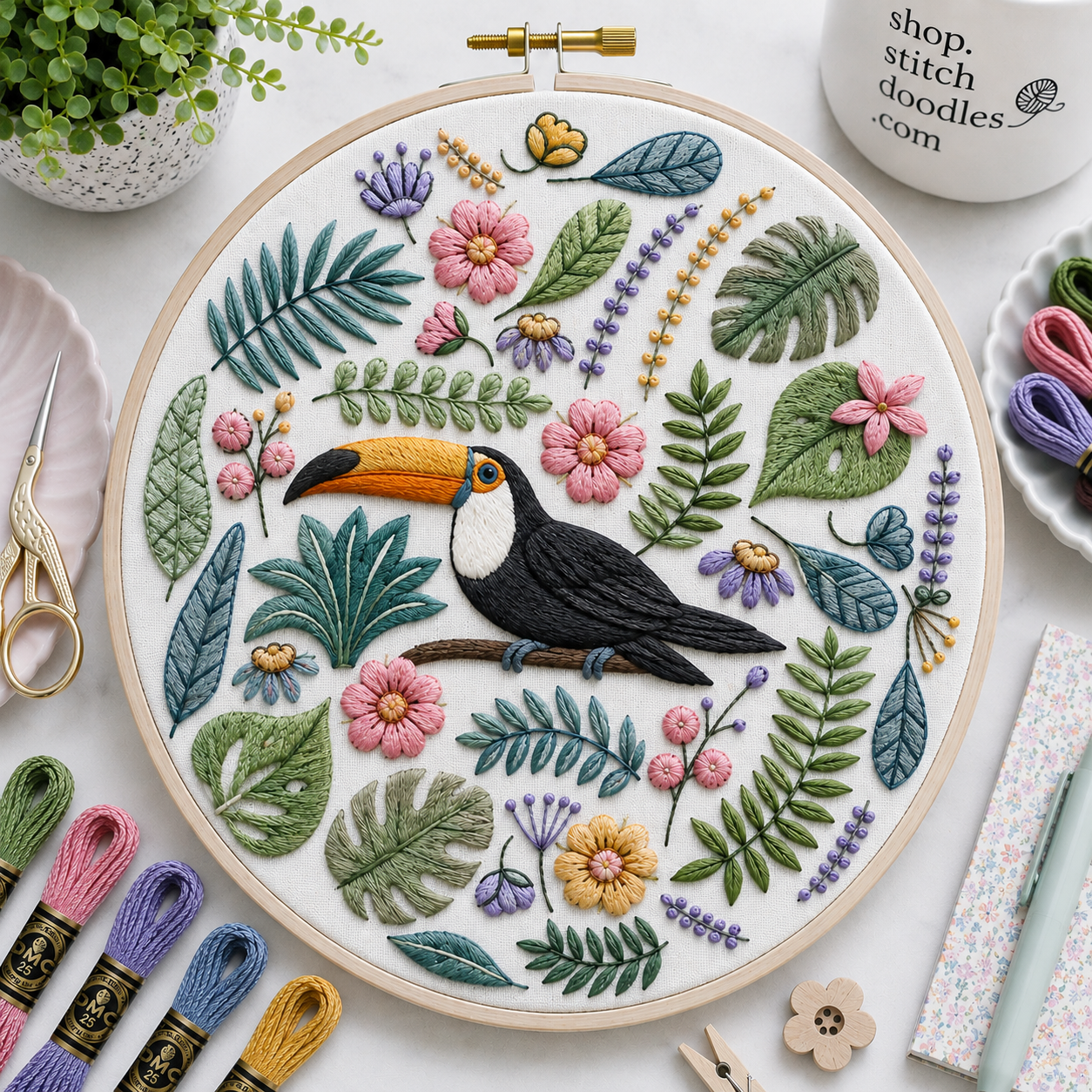 Embroidery hoop with a toucan design surrounded by flowers and leaves on a table with embroidery supplies.
