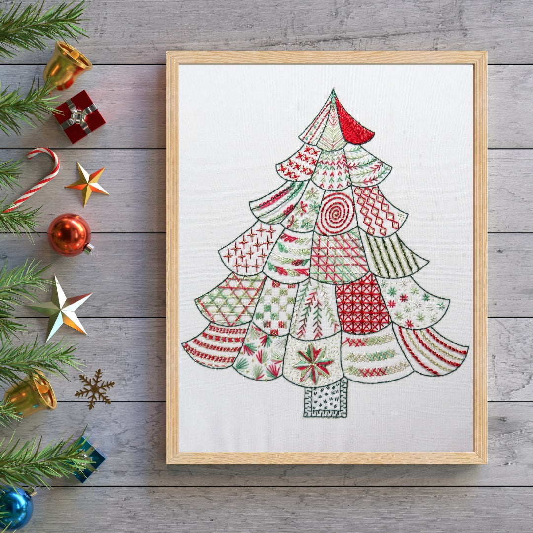 A framed art print featuring a stylized Christmas tree made of layered patterned fabric-like shapes in red, green, and cream, displayed on a gray wood plank surface with holiday decorations around it.