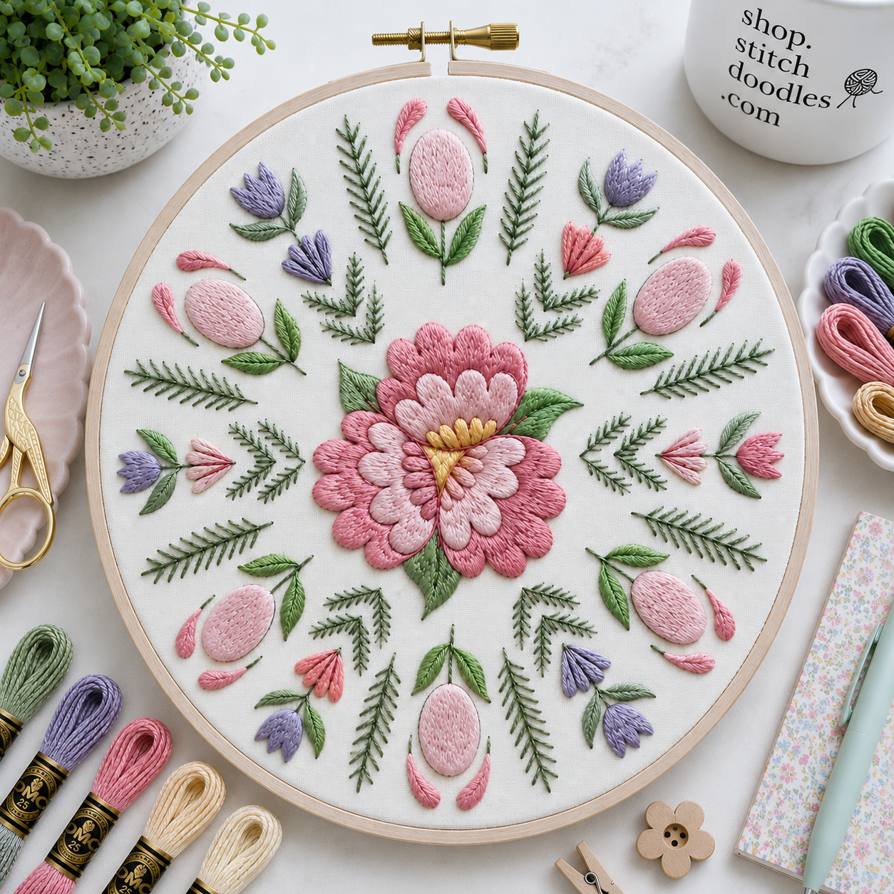 Floral embroidery hoop with pink and green flowers on a white background, surrounded by embroidery supplies.