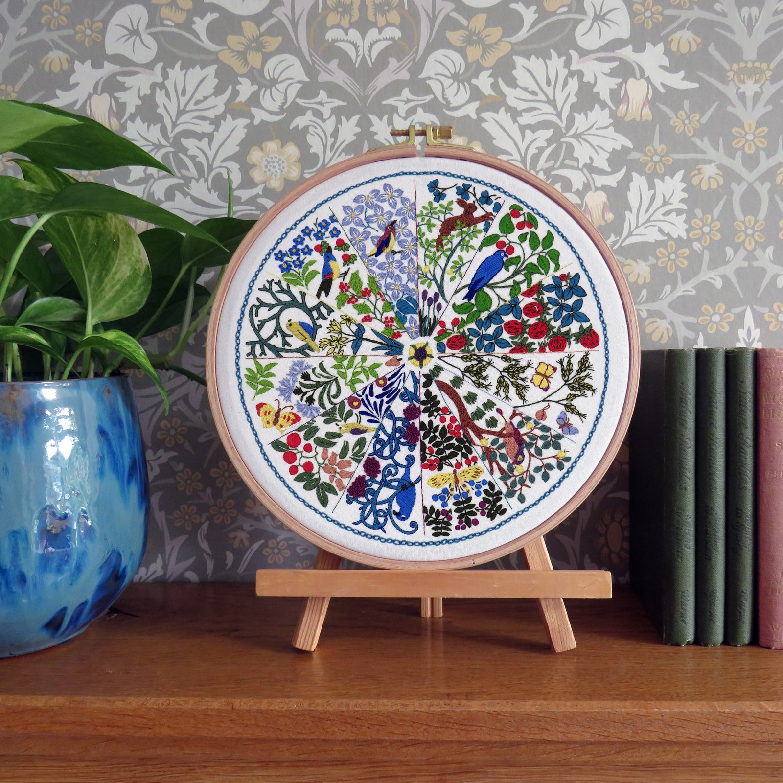 Embroidery hoop with floral and bird design on a wooden stand against a decorative wall.