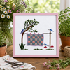 Embroidery of a garden scene with a tree, birds, and a checkered cloth, framed in pink.