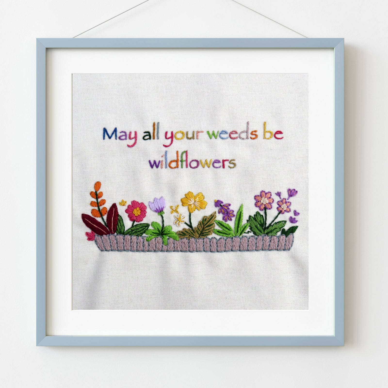 Embroidered artwork with flowers and text 'May all your weeds be wildflowers' in a light blue frame.