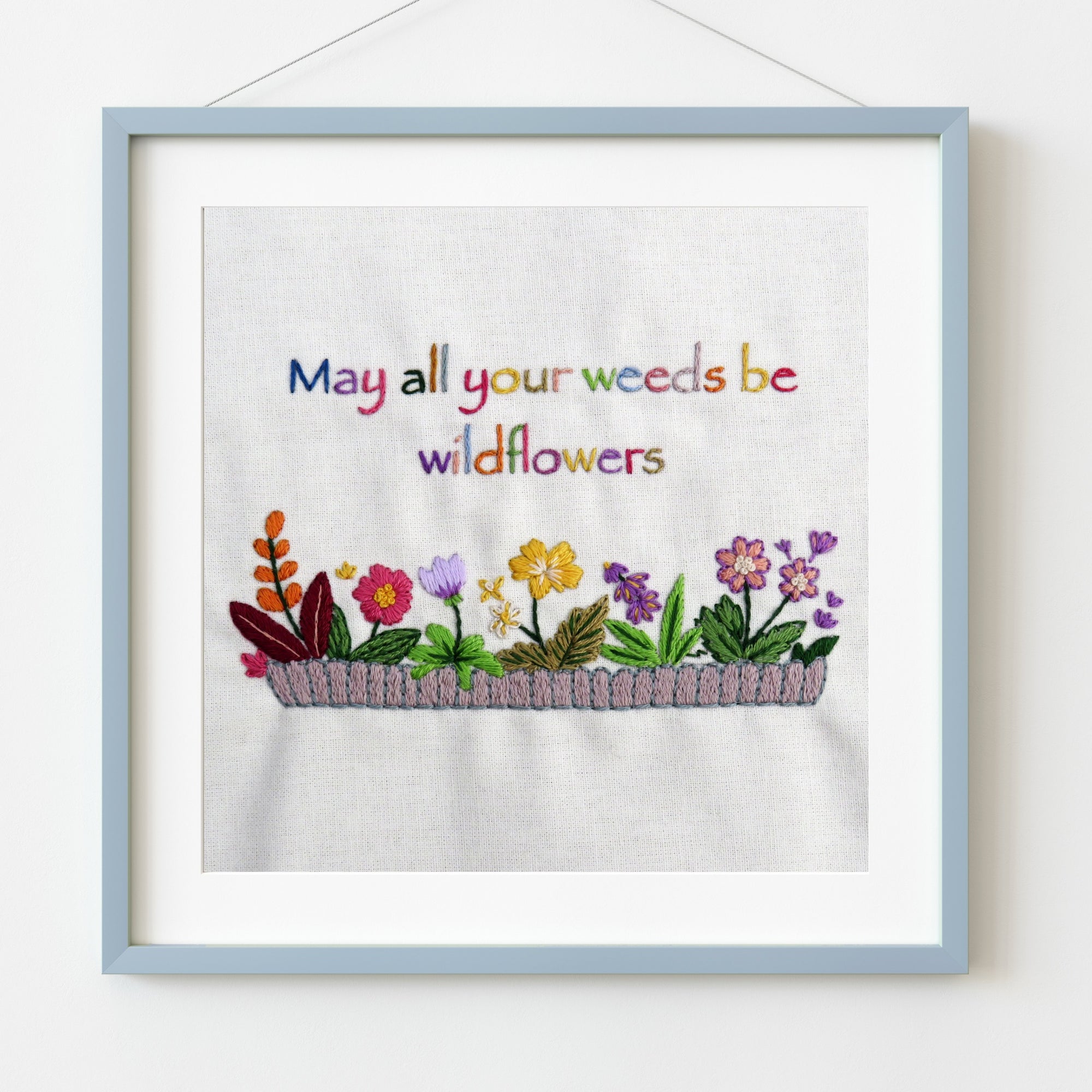 Embroidered artwork with flowers and text 'May all your weeds be wildflowers' in a light blue frame.