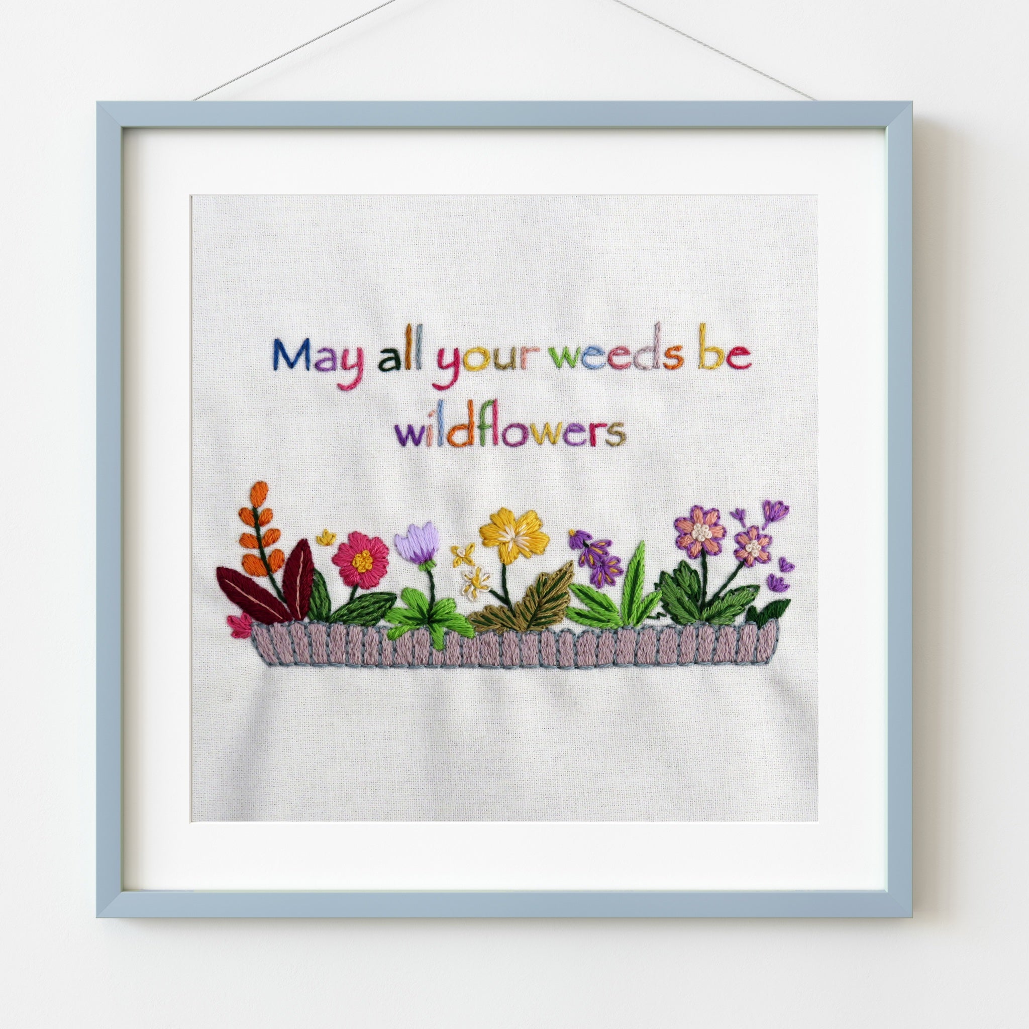 Embroidered artwork with flowers and text 'May all your weeds be wildflowers' in a light blue frame.