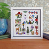 A framed square embroidery sampler featuring whimsical woodland and garden motifs, displayed on a wooden surface against patterned wallpaper with a potted plant and book nearby.