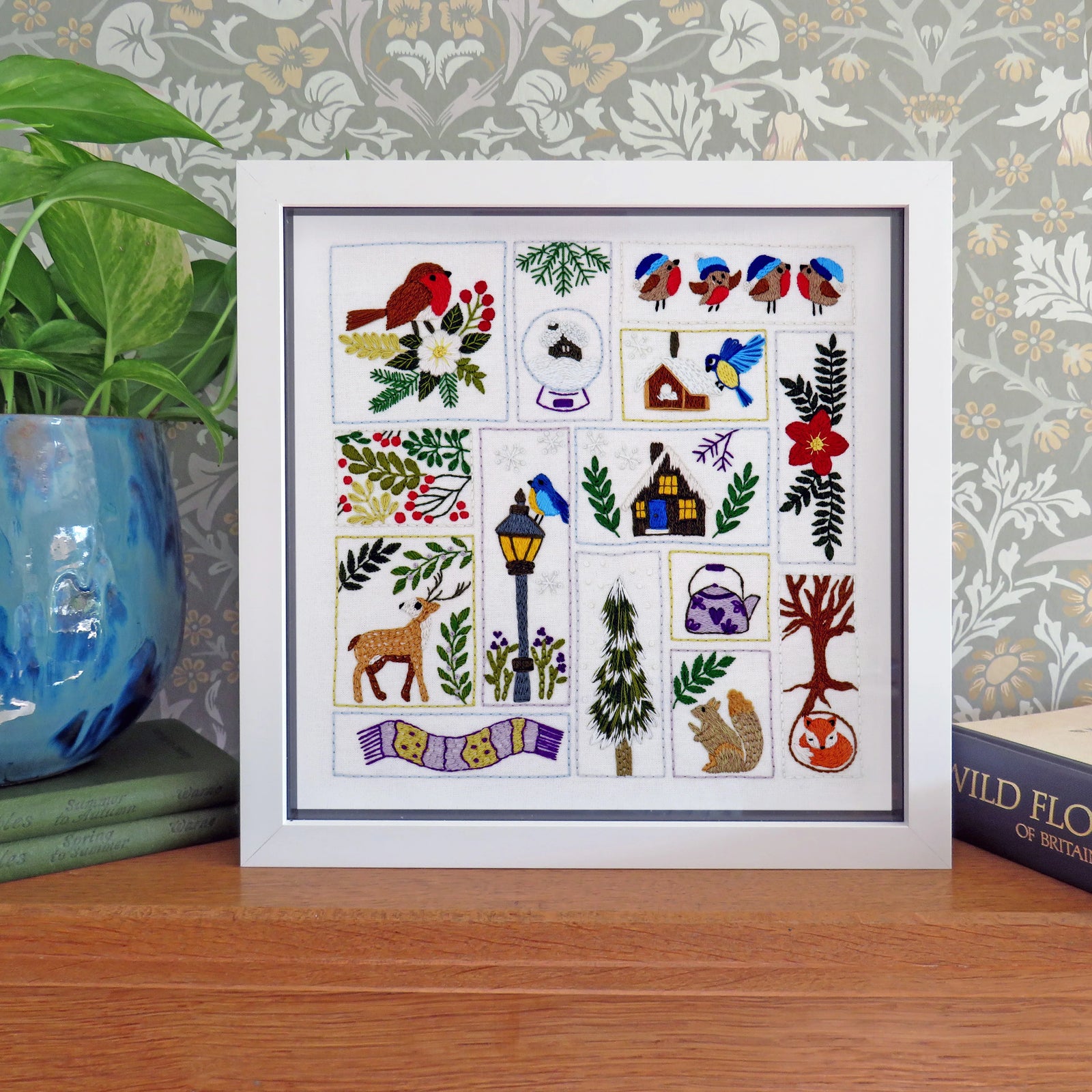 A framed square embroidery sampler featuring whimsical woodland and garden motifs, displayed on a wooden surface against patterned wallpaper with a potted plant and book nearby.