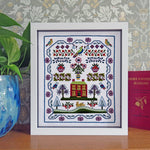 A framed completed cross-stitch or embroidery piece displayed on a wooden surface, featuring a colorful cottage, birds, flowers, and trees. The white-framed artwork is set against patterned wallpaper, with a blue glass vase on the left and a red book on the right.
