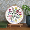 A circular embroidery hoop displaying a colorful floral botanical design, mounted on a small wooden stand on a wooden table. The background features patterned wallpaper, a green potted plant, and stacked books.