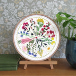 A circular embroidery hoop displaying a colorful floral botanical design, mounted on a small wooden stand on a wooden table. The background features patterned wallpaper, a green potted plant, and stacked books.
