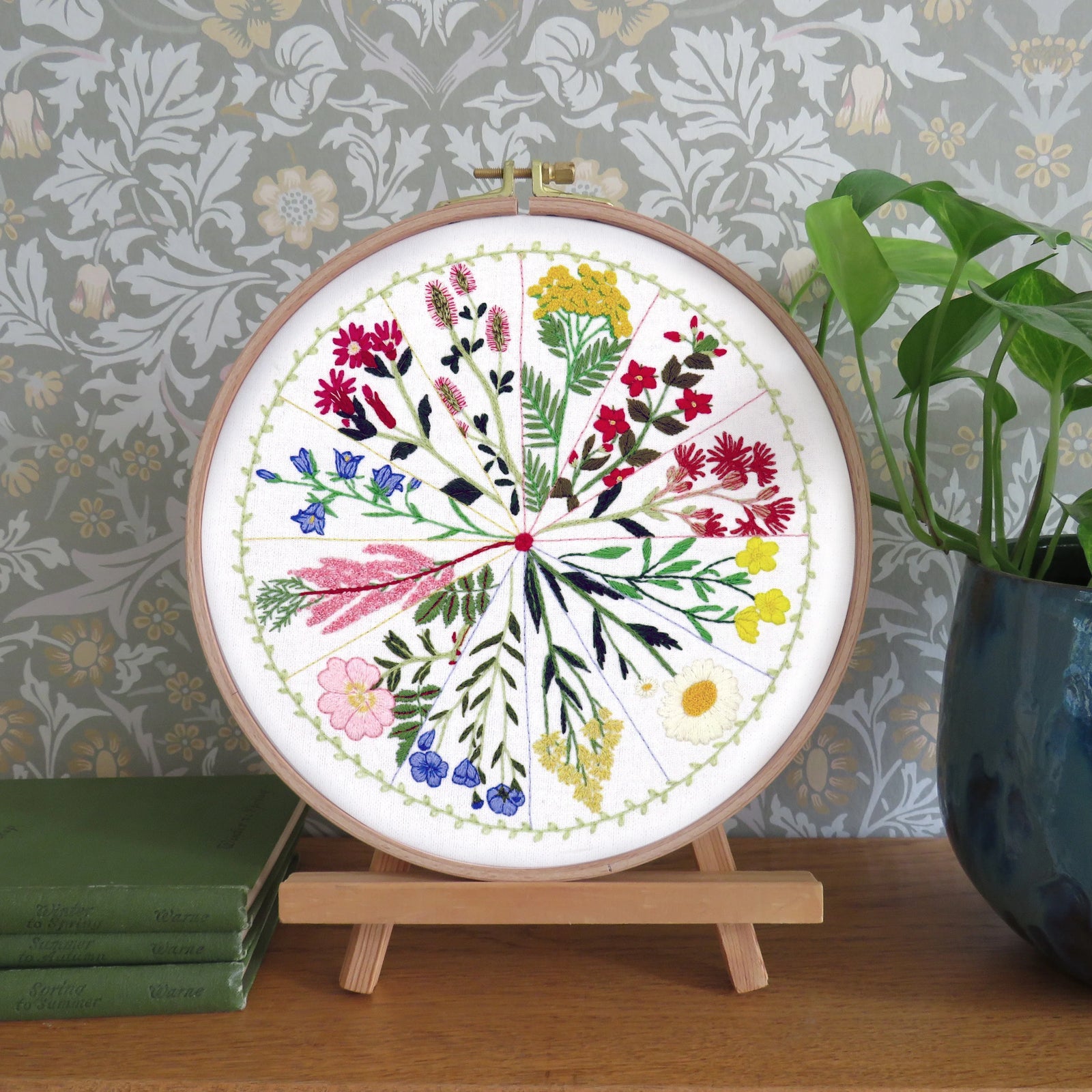 A circular embroidery hoop displaying a colorful floral botanical design, mounted on a small wooden stand on a wooden table. The background features patterned wallpaper, a green potted plant, and stacked books.