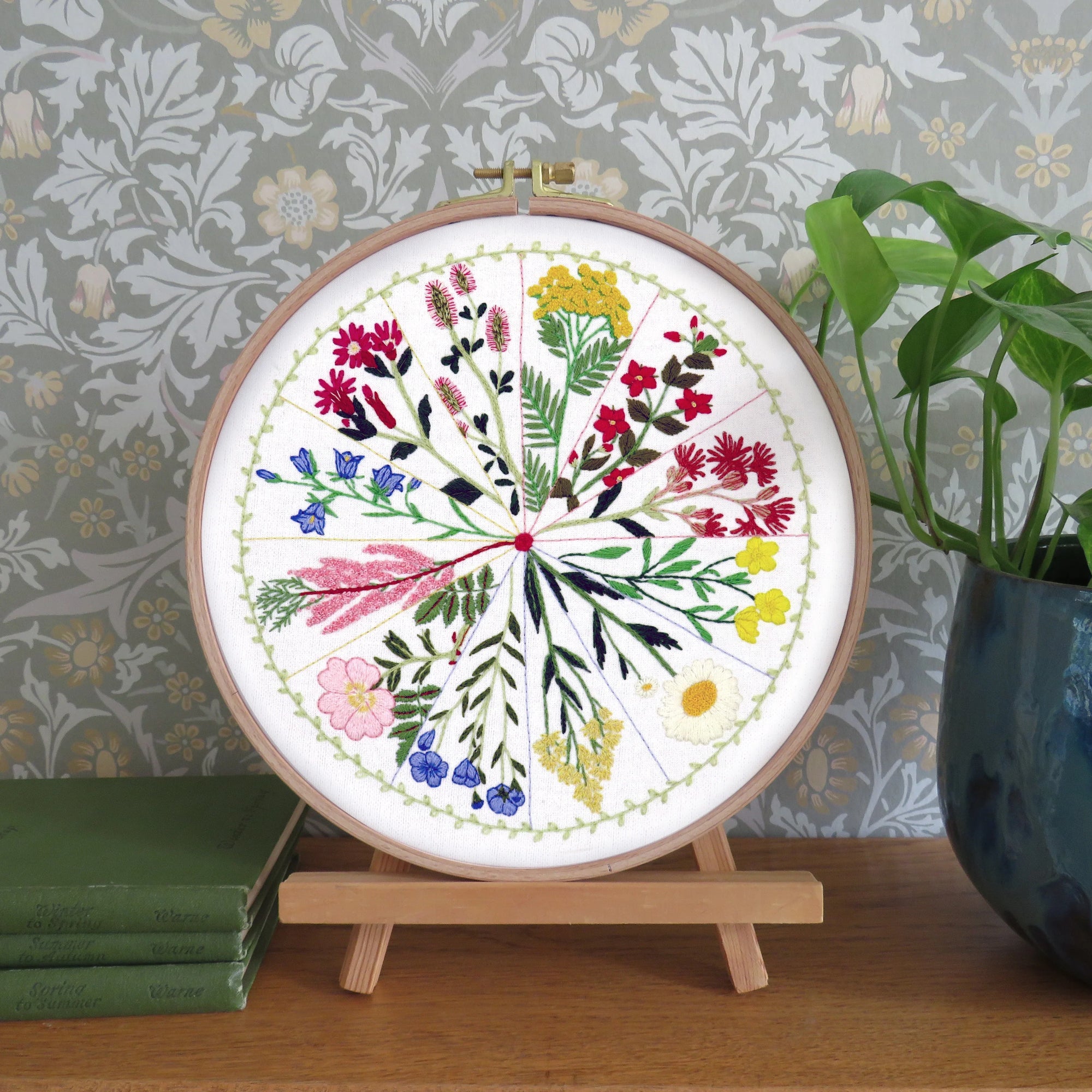 A circular embroidery hoop displaying a colorful floral botanical design, mounted on a small wooden stand on a wooden table. The background features patterned wallpaper, a green potted plant, and stacked books.