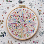 A round wooden embroidery hoop holding a colorful monthly hand embroidery cross-stitch chart with illustrated seasonal motifs and month labels, placed on a matching fabric background with botanical illustrations and seasonal words.