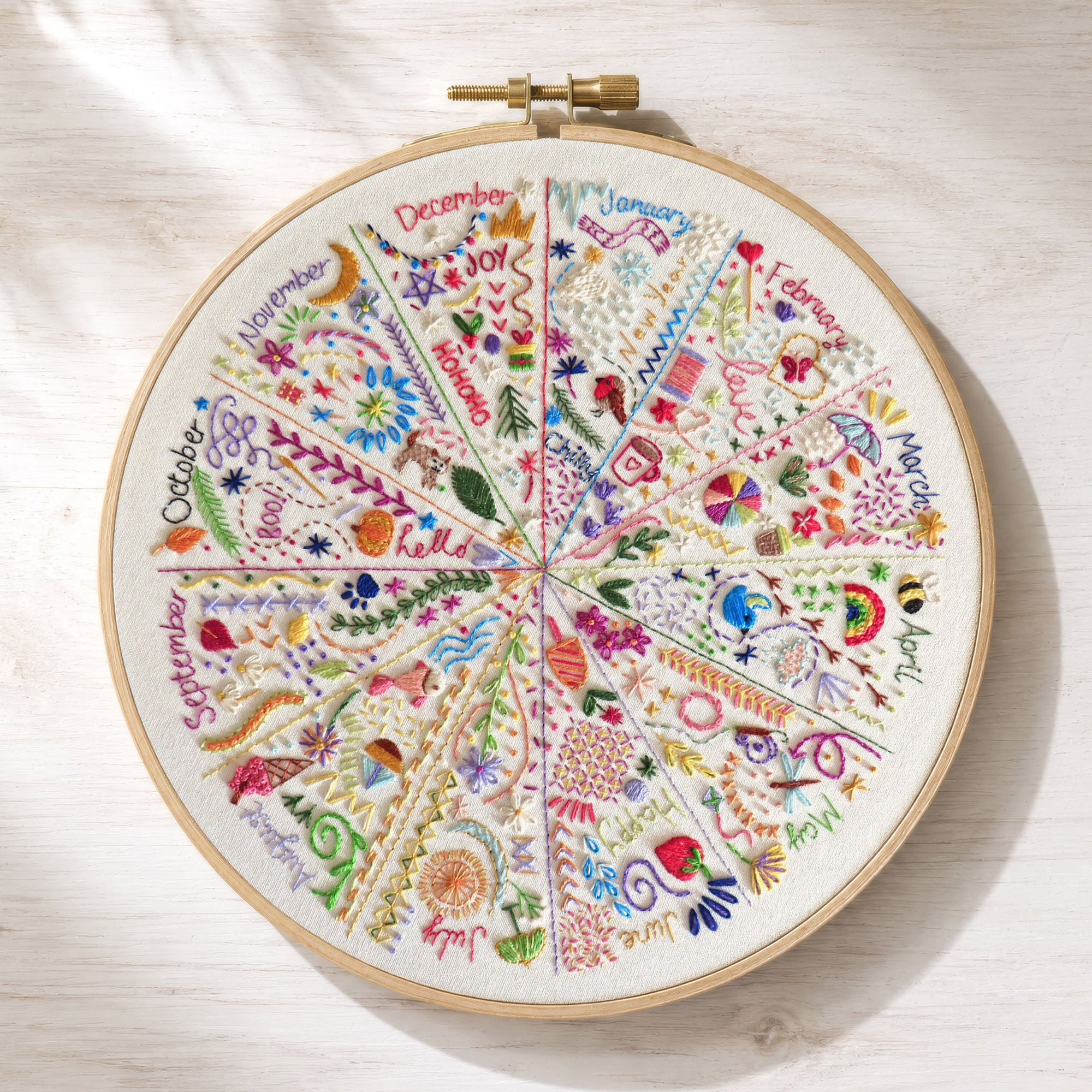 A circular wooden embroidery hoop containing a colorful monthly floral and botanical chart, displayed on a light wood surface with soft leaf shadows.