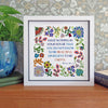 A framed William Morris cross-stitch style print featuring the quote 'HAVE NOTHING IN YOUR HOUSE THAT YOU DO NOT KNOW IS BEAUTIFUL OR BELIEVE TO BE USEFUL' surrounded by colorful floral motifs, displayed on a wooden surface next to a potted plant.