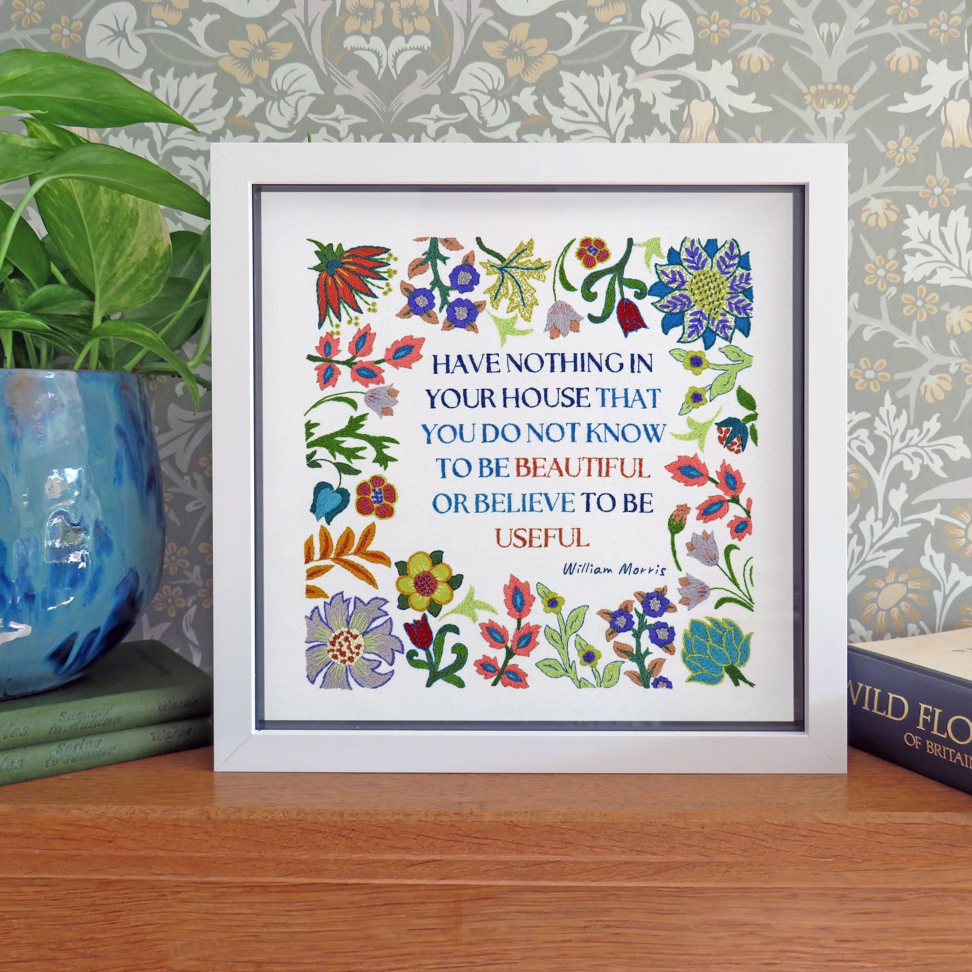A framed William Morris cross-stitch style print featuring the quote 'HAVE NOTHING IN YOUR HOUSE THAT YOU DO NOT KNOW IS BEAUTIFUL OR BELIEVE TO BE USEFUL' surrounded by colorful floral motifs, displayed on a wooden surface next to a potted plant.